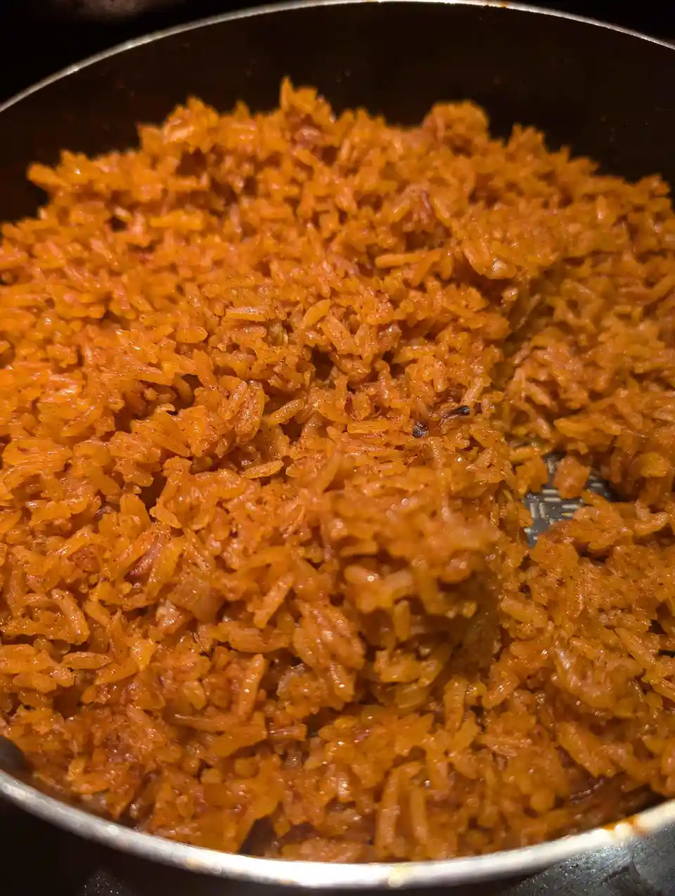 A close-up photo shows a pan filled with vibrant orange-colored rice, likely seasoned with spices, with a portion scooped…