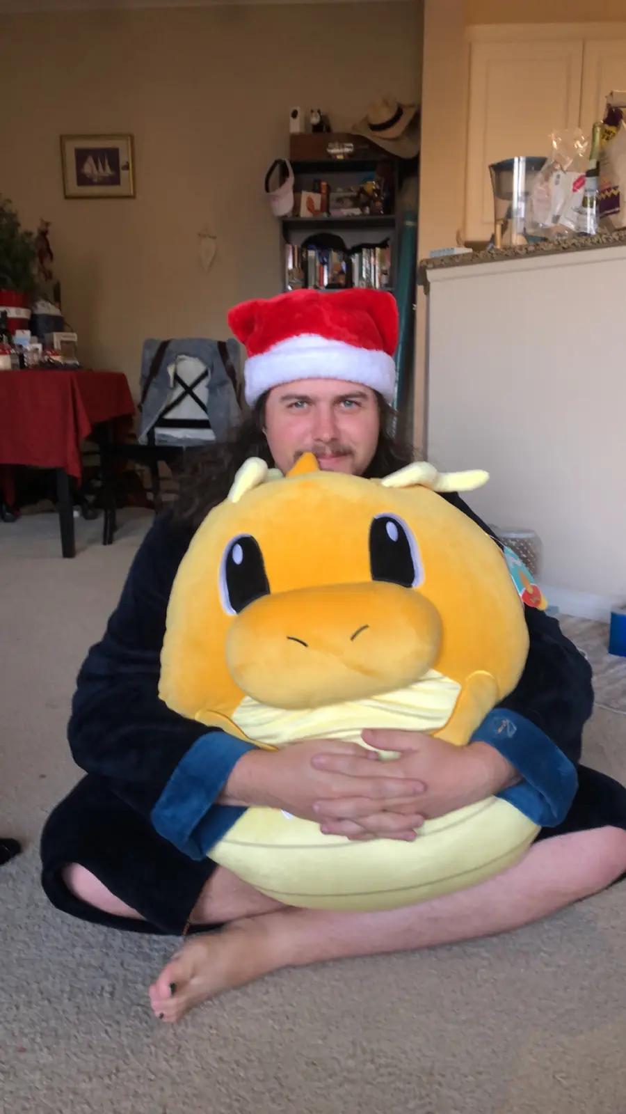 A man with long hair and a mustache, wearing a red and white Santa hat and dark blue pajamas, sits cross-legged on a…