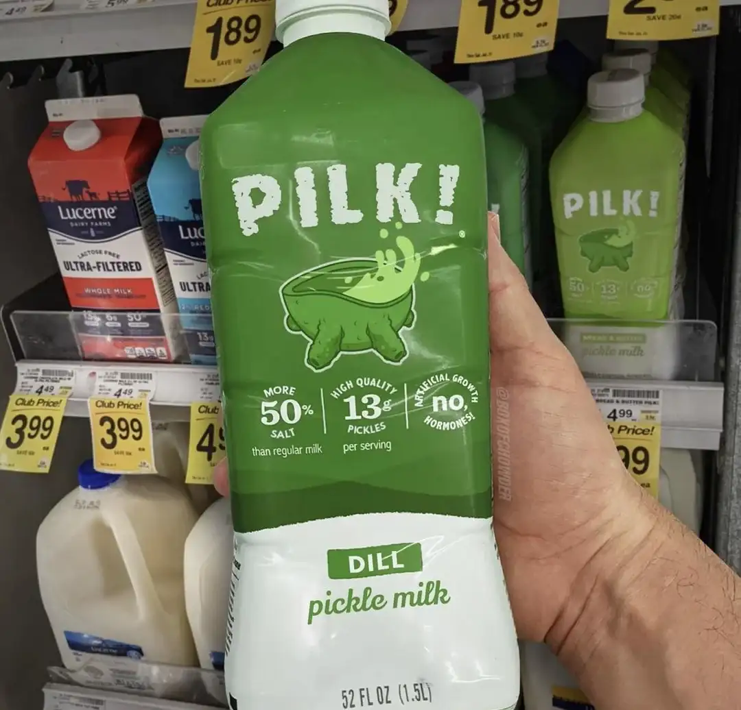 A hand holds a large green bottle of 'PILK! DILL pickle milk' in a grocery store dairy aisle, surrounded by regular milk…