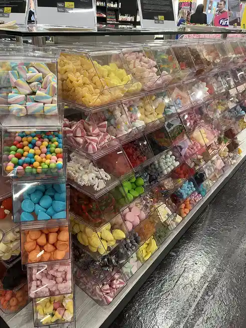A photo shows a long, multi-tiered display case filled with numerous clear bins of assorted colorful candies like gummy…