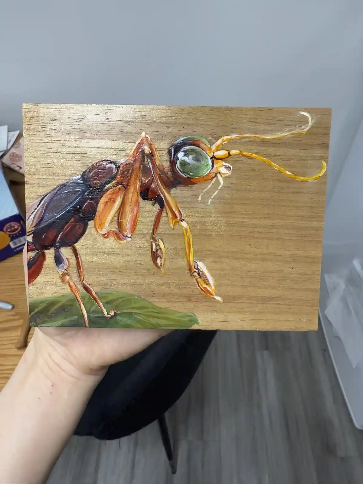 A hand holds up a small wooden painting depicting a highly detailed, realistic insect with large green eyes, amber legs, and…