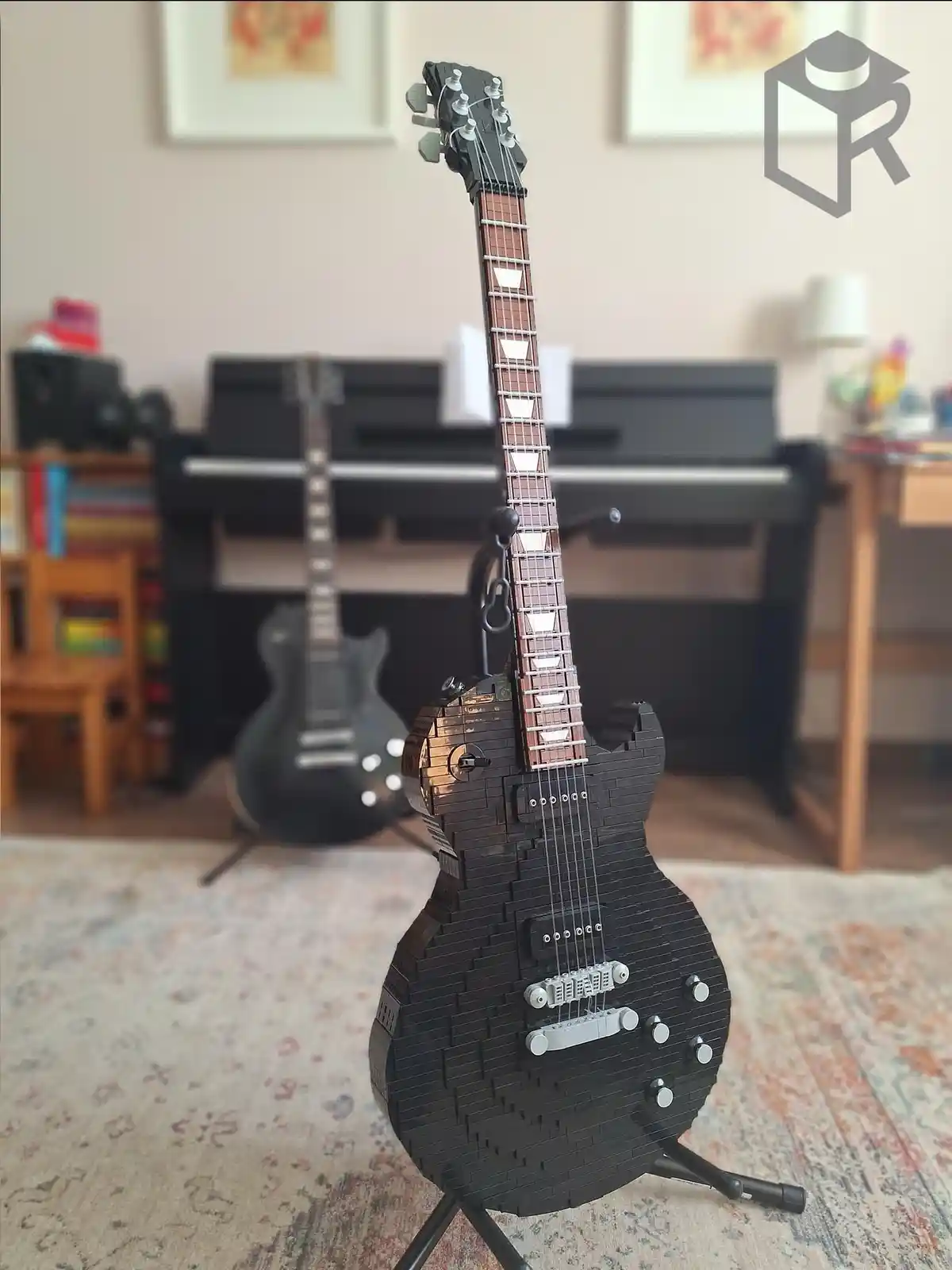 A black electric guitar meticulously constructed from LEGO bricks stands upright on a stand in a room, with a real black…