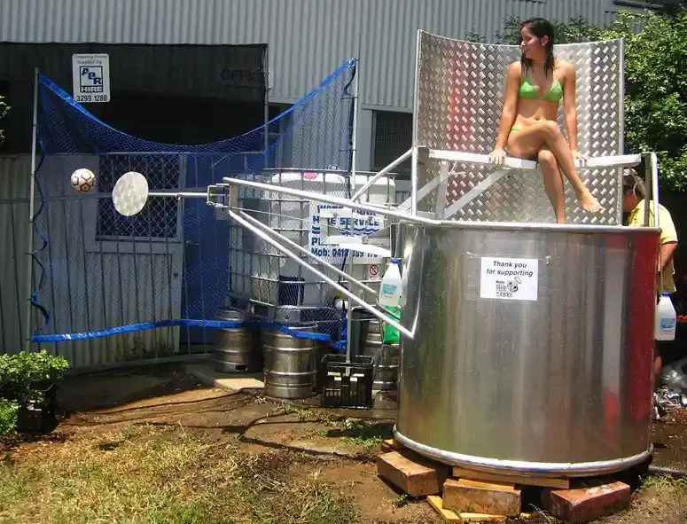 A woman in a green bikini sits atop a large metal beer tank outdoors, with a soccer ball on a pole nearby and a blue netted…