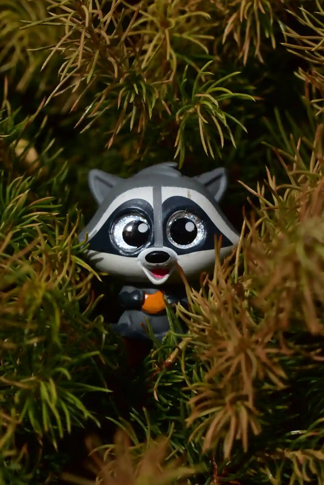 A small, stylized raccoon toy with large, glittery eyes and a surprised expression is partially hidden among green and brown…