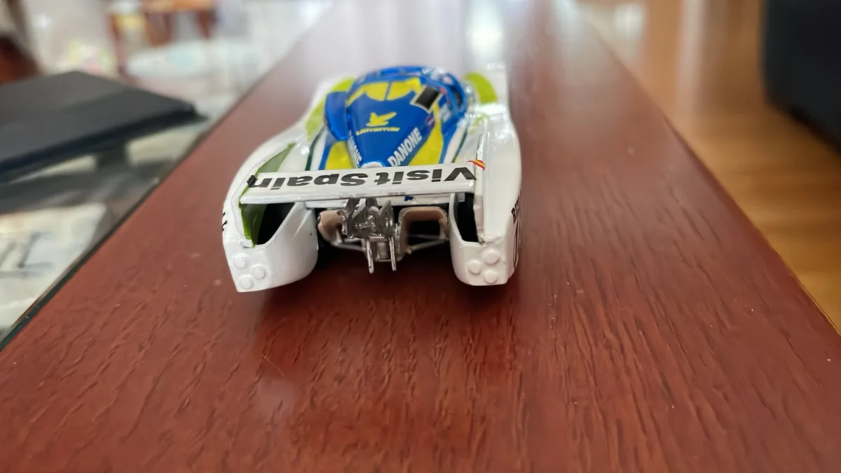 A close-up photo of a white, blue, and yellow toy race car model viewed from the rear, sitting on a wooden table. The car's…
