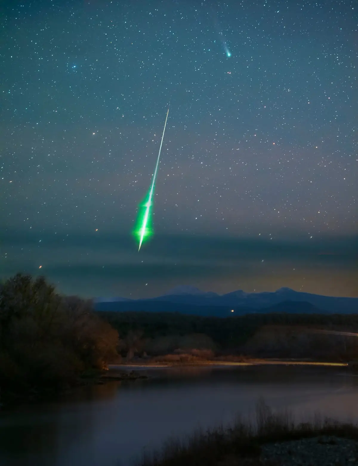 A long-exposure night photograph shows a starry sky over a calm lake and distant mountains, with a bright green comet…