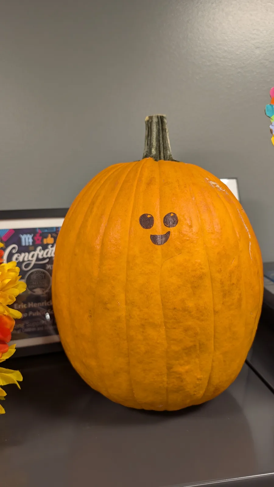 A large, orange pumpkin with a simple, hand-drawn smiling face—two round eyes and a curved mouth—is sitting on a dark…