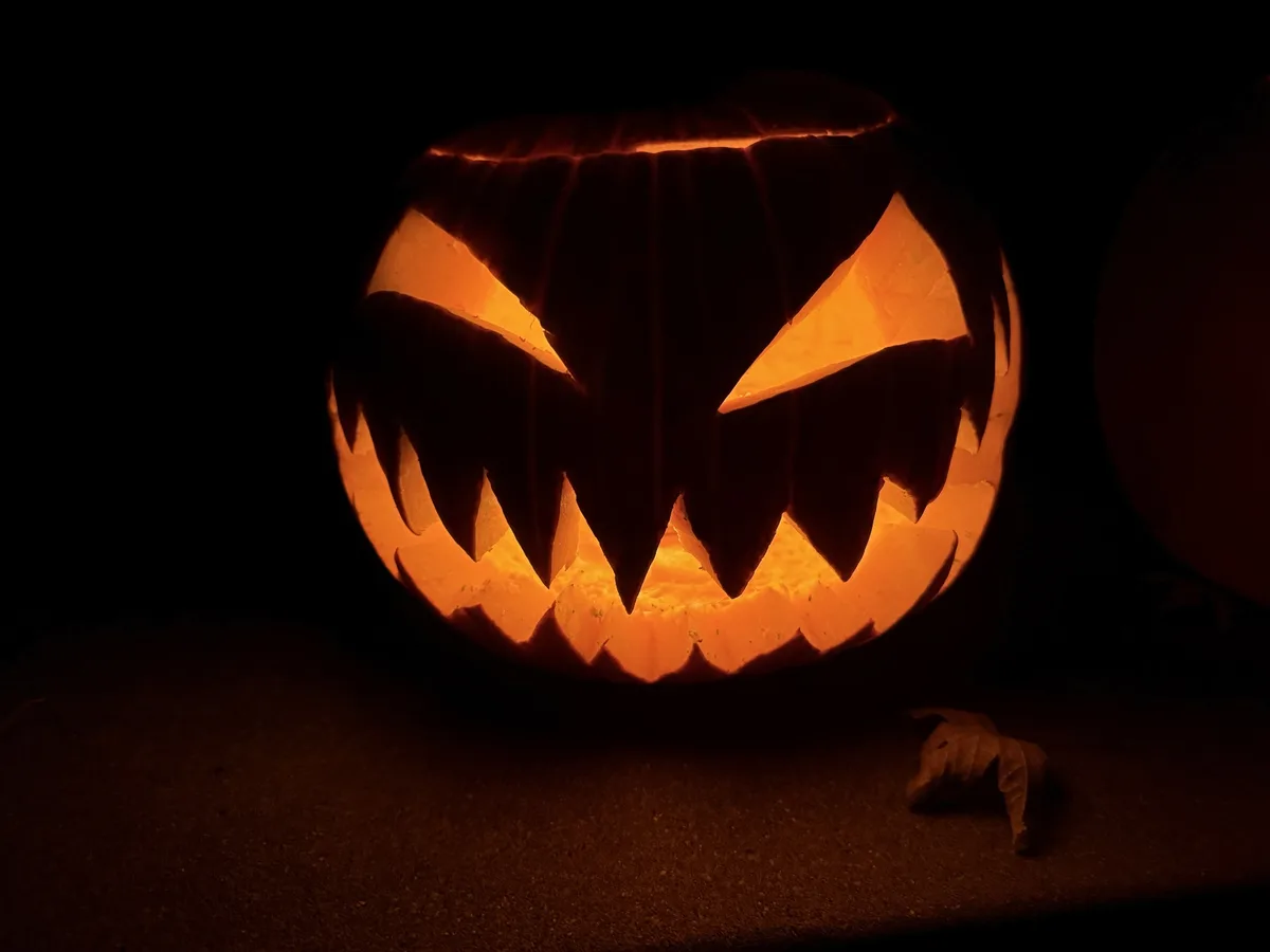 A carved pumpkin with glowing orange eyes and jagged teeth sits in darkness, illuminated from within, with a dry leaf…