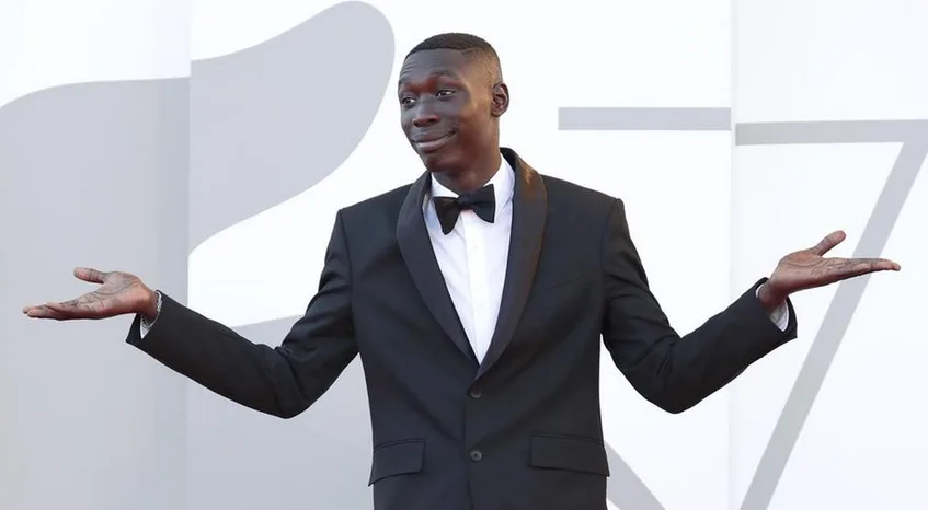 a man in a black tuxedo with a bow tie stands with arms outstretched palms up, looking confused or exasperated, against a…