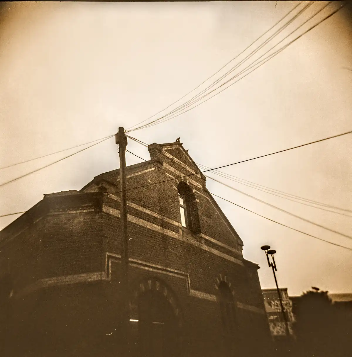 A sepia-toned, low-angle photograph of a brick building with a gabled roof, power lines crisscrossing the sky, and a utility…