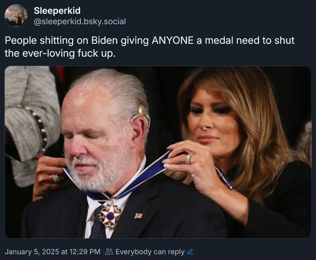A man with a white beard and hearing aid is having a medal with a blue ribbon pinned around his neck by a woman with long…