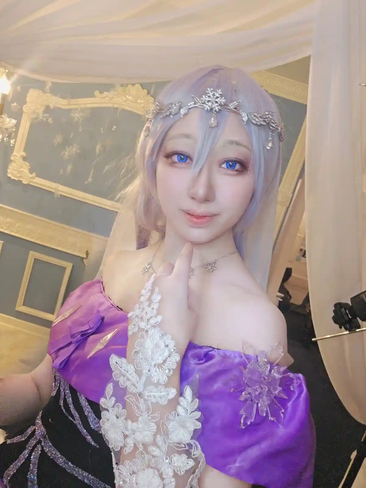 A person with light purple hair and striking blue eyes poses in a detailed costume featuring a purple off-the-shoulder top…