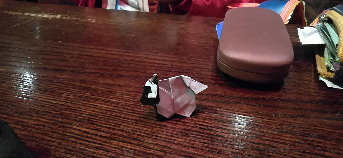 A small origami cow, made from pink and black paper, sits on a dark wooden table. Nearby are a brown glasses case and a…