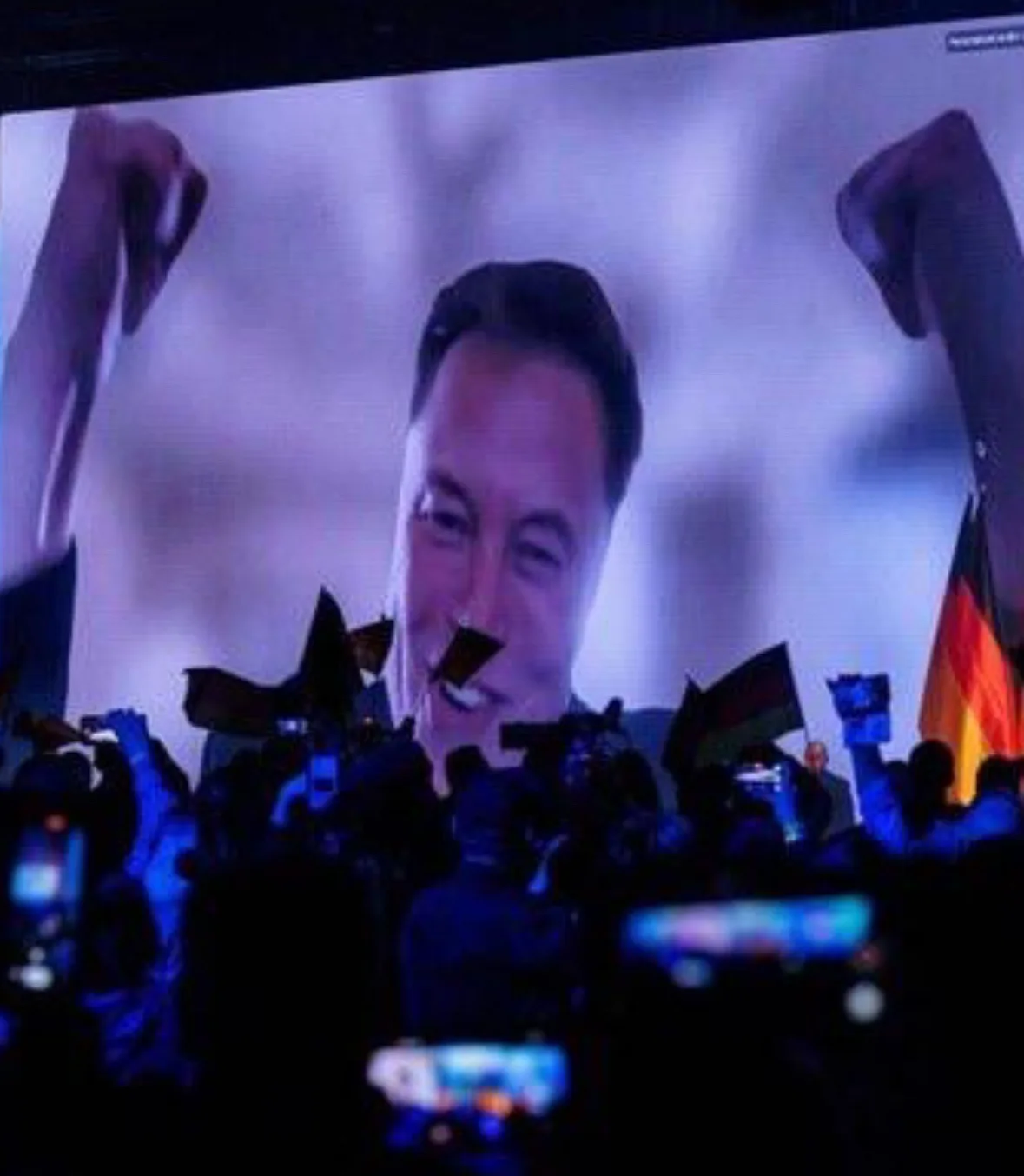 A large screen displays a smiling man with both fists raised in the air, likely Elon Musk, while below, a crowd of people…