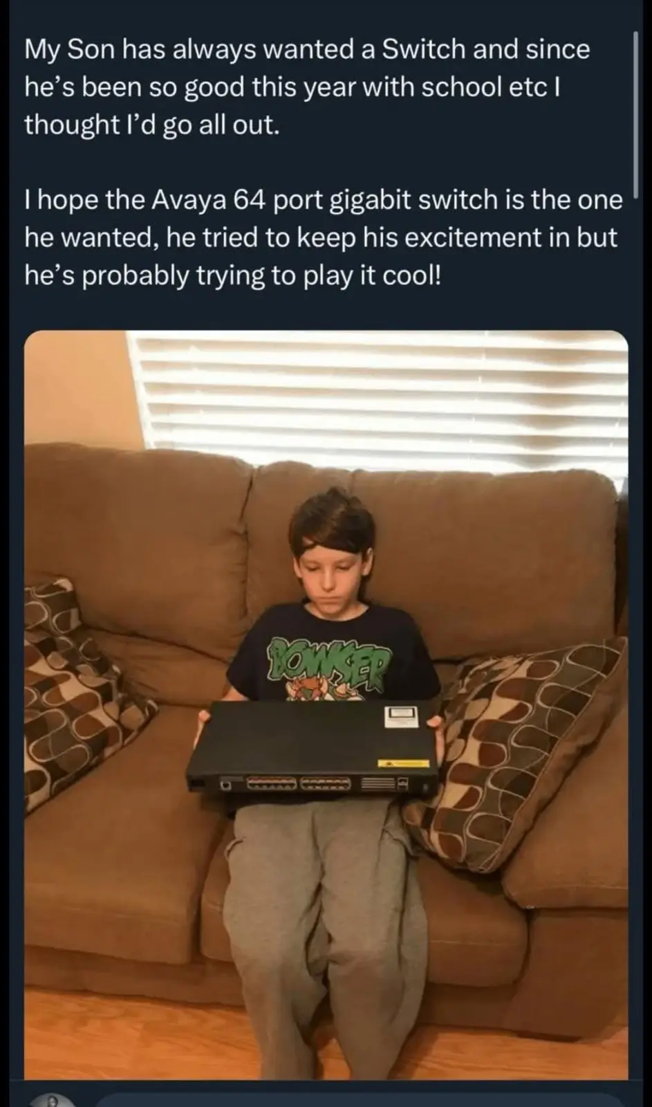 A young boy sits on a brown couch holding a large, black, professional-grade Avaya 64-port gigabit network switch, looking…