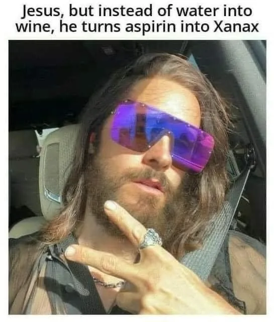 A man with long hair and a beard wearing large purple-tinted sunglasses sits in a car, making a peace sign with his hand. He…