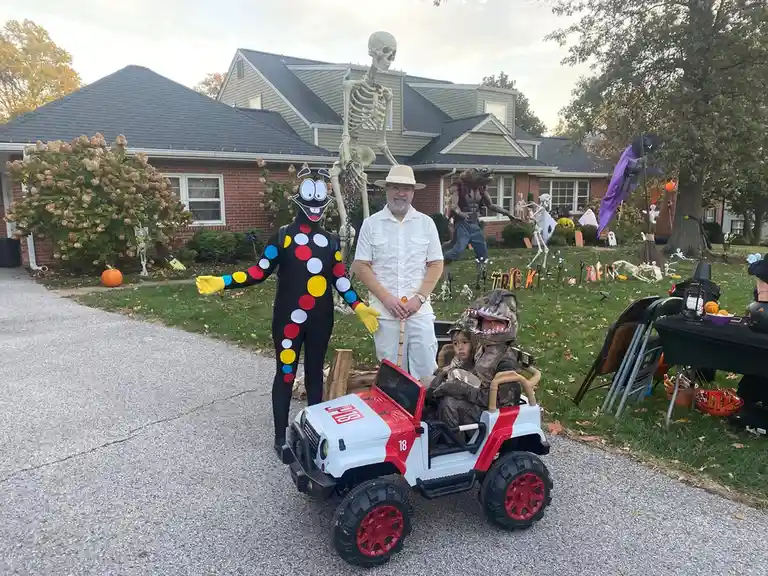 A man in a white outfit stands beside a child in a dinosaur costume riding a red and white toy jeep, next to a person in a…