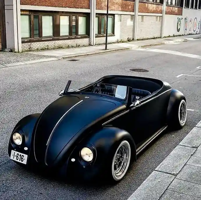 A matte black, custom lowrider Volkswagen Beetle convertible is parked on an asphalt street next to a sidewalk, with a brick…