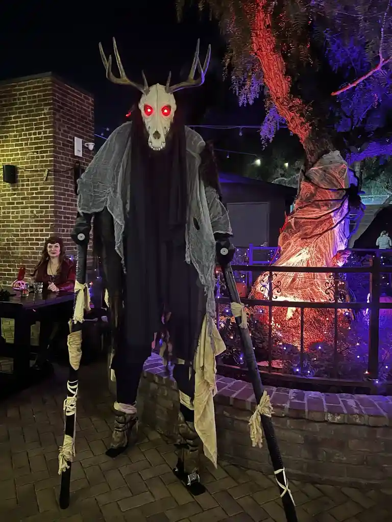 A tall, eerie Halloween figure with a deer skull mask featuring glowing red eyes, antlers, and a tattered grey cloak stands…