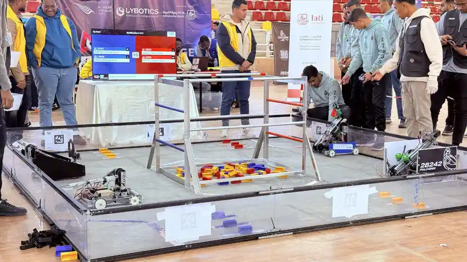 A robotics competition scene with two robotic combatants in a glass arena, surrounded by team members and spectators. One…
