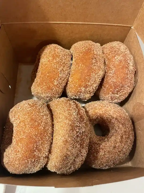 A top-down view of a cardboard box containing six sugar-coated donuts arranged to visually spell out the number '100'…