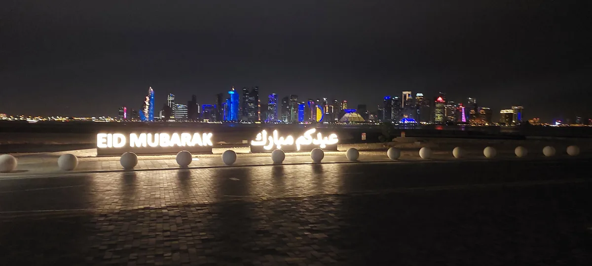 A nighttime photo of a city skyline with illuminated skyscrapers, featuring two large glowing signs in the foreground: one…