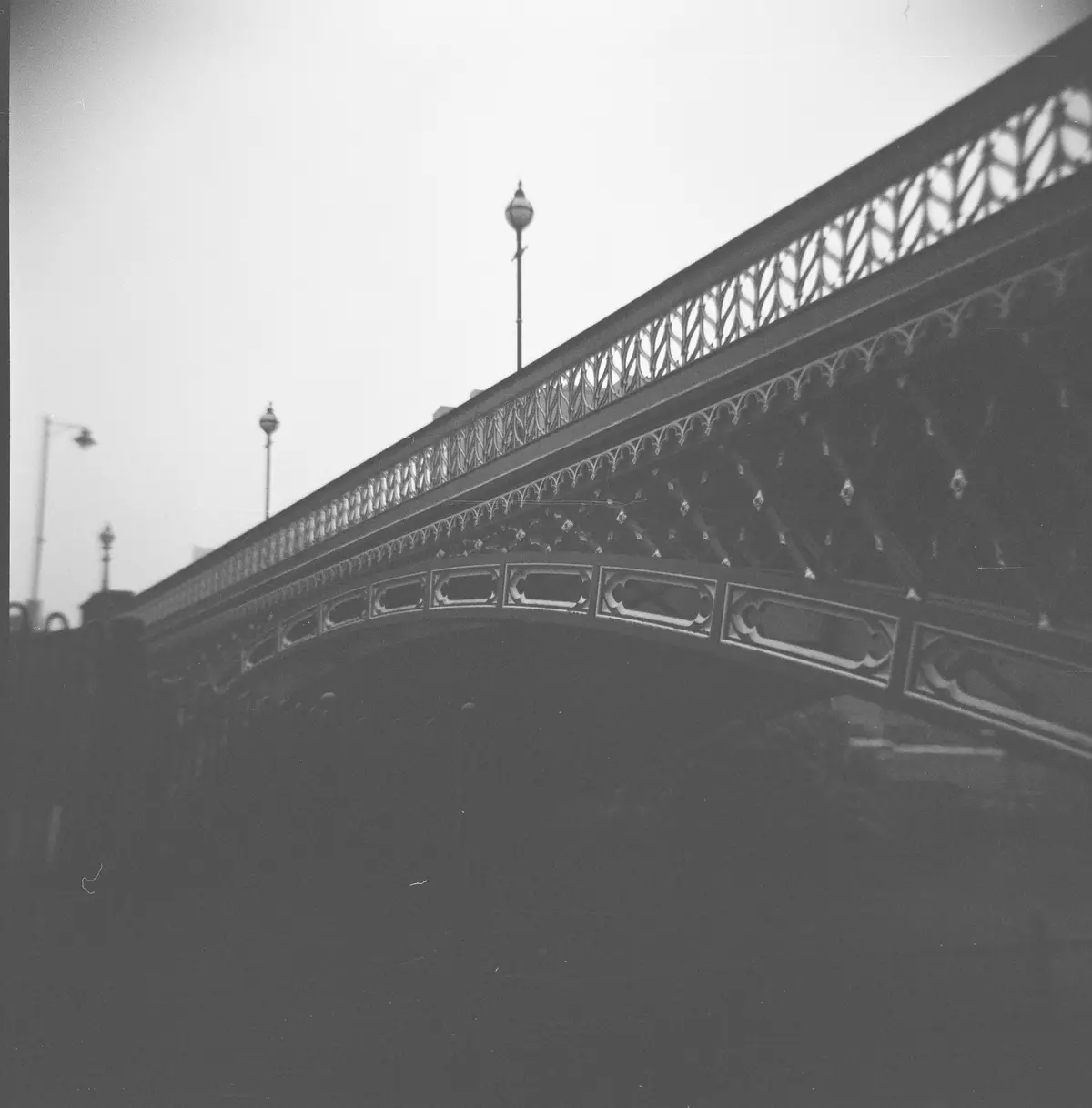 A black-and-white photograph shows the ornate, arched metalwork of a bridge, likely the Iron Bridge, viewed from below with…