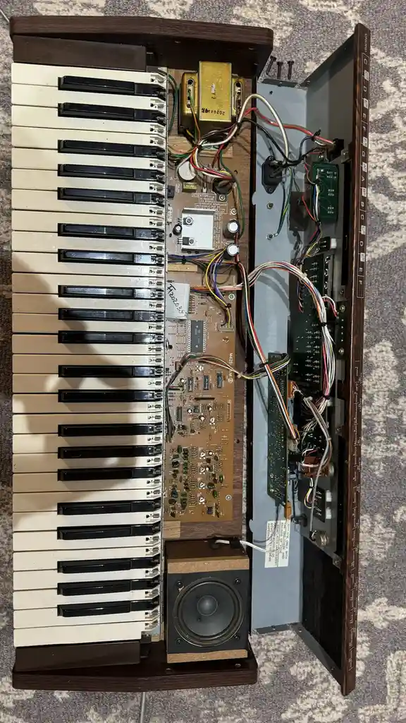 An overhead photo shows a vintage wooden keyboard instrument with its casing removed, exposing a complex mess of circuit…