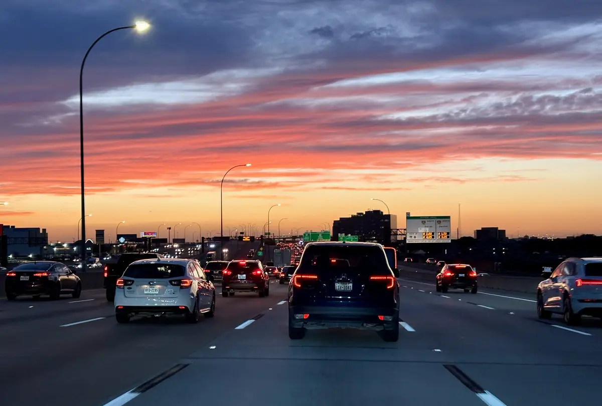 A photo of heavy traffic on a highway at sunset, with cars' brake lights glowing red against a vibrant orange and purple…