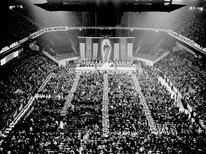 A black-and-white photo shows a massive crowd in a large arena, with rows of people standing in formation leading to a…