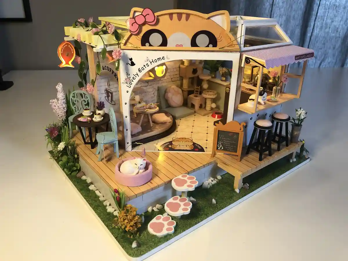 A detailed miniature model of a cat café, featuring a cat-shaped roof with large eyes and a pink bow, a front patio with a…