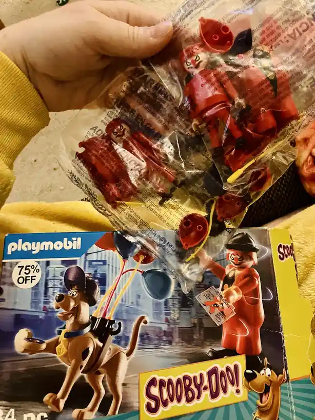A hand holds a Playmobil Scooby-Doo toy package, showing a red clown figure inside the clear plastic. The box art features…