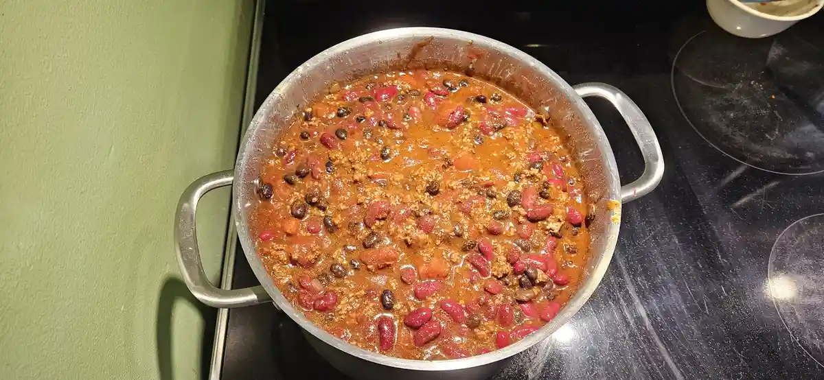 A large stainless steel pot sits on a black stovetop, filled with a thick, reddish-brown chili containing visible red kidney…