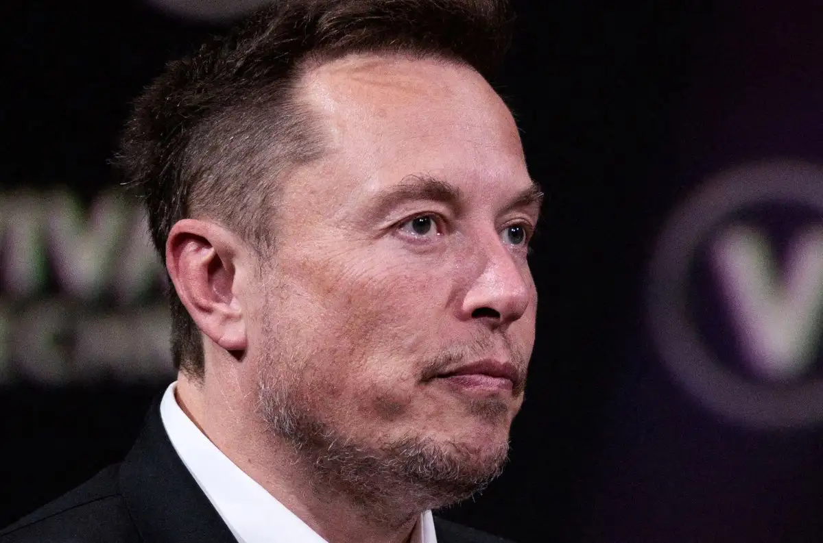 a close-up photo of elon musk looking slightly to his right with a neutral expression, wearing a black suit and white shirt…