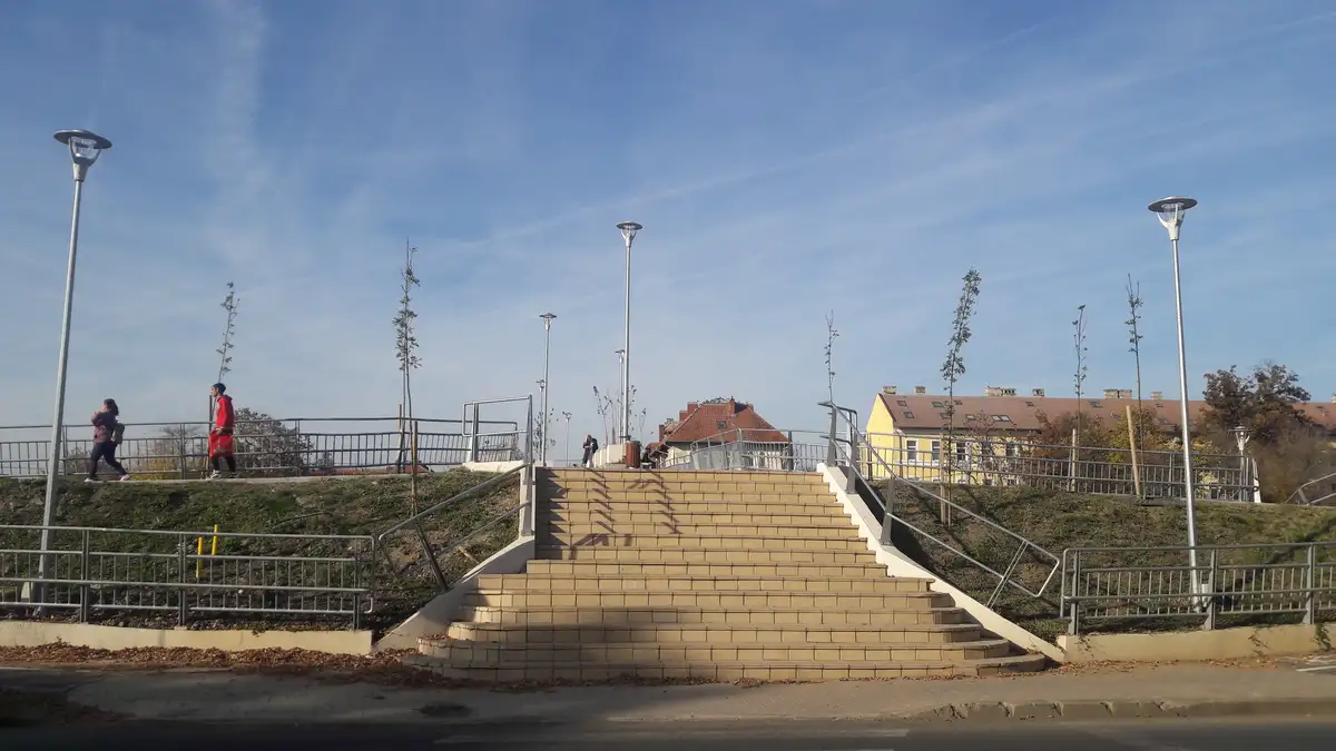 A wide outdoor staircase with metal railings and streetlights under a clear blue sky, with a few people walking on the upper…