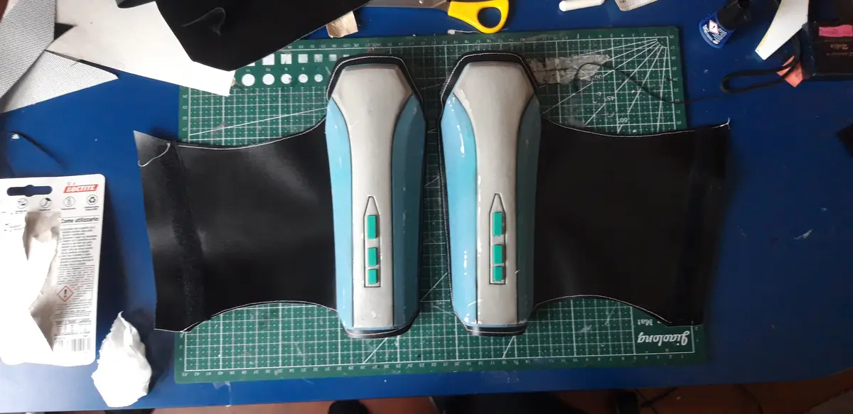 Two custom-made, light blue and silver cosplay gauntlets with green rectangular details sit on a green cutting mat, flanked…