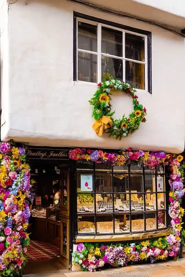 A jeweler's storefront is extravagantly decorated with a dense, colorful garland of artificial flowers framing the windows…