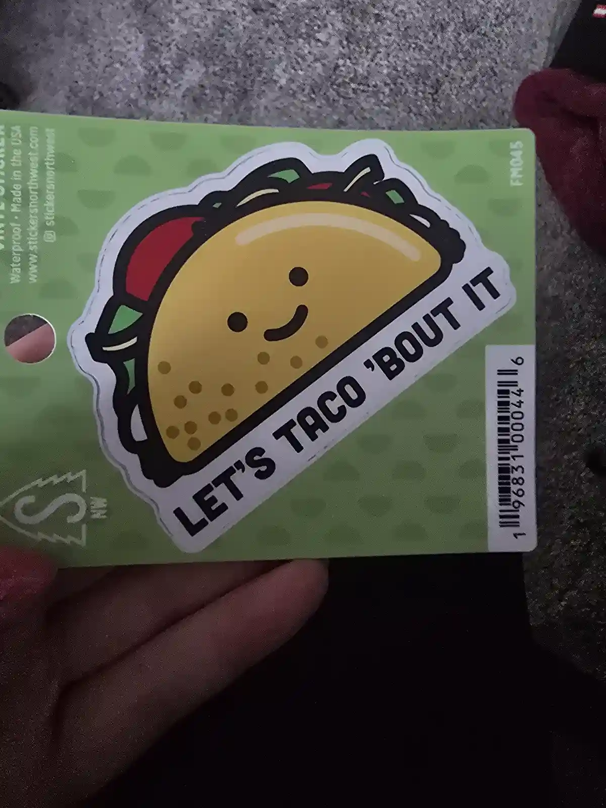 A hand holds a green sticker sheet featuring a cartoon taco with a smiling face and the text 'LET'S TACO 'BOUT IT' along its…