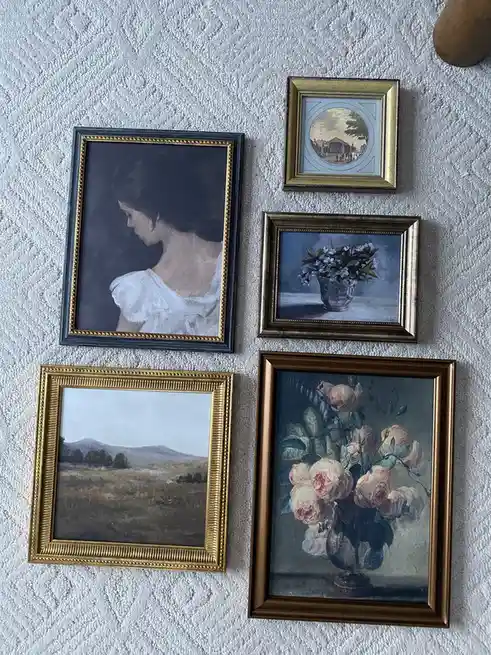 Five framed paintings hang on a textured white wall, including a portrait of a woman in profile, a still life of flowers in…