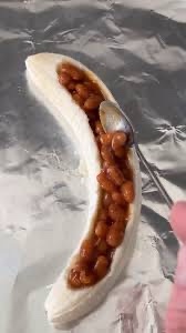 A peeled banana lies on aluminum foil, filled with baked beans, with a spoon inserted into the beans. A hand holds the…