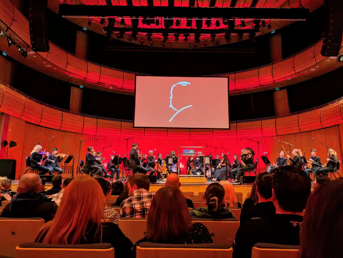 An orchestra performs on a stage in a concert hall with red lighting, while a large screen behind them displays a minimalist…
