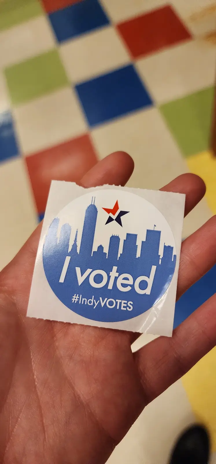 A hand holds a circular blue and white 'I voted' sticker featuring a city skyline and a star, with the hashtag #IndyVOTES…