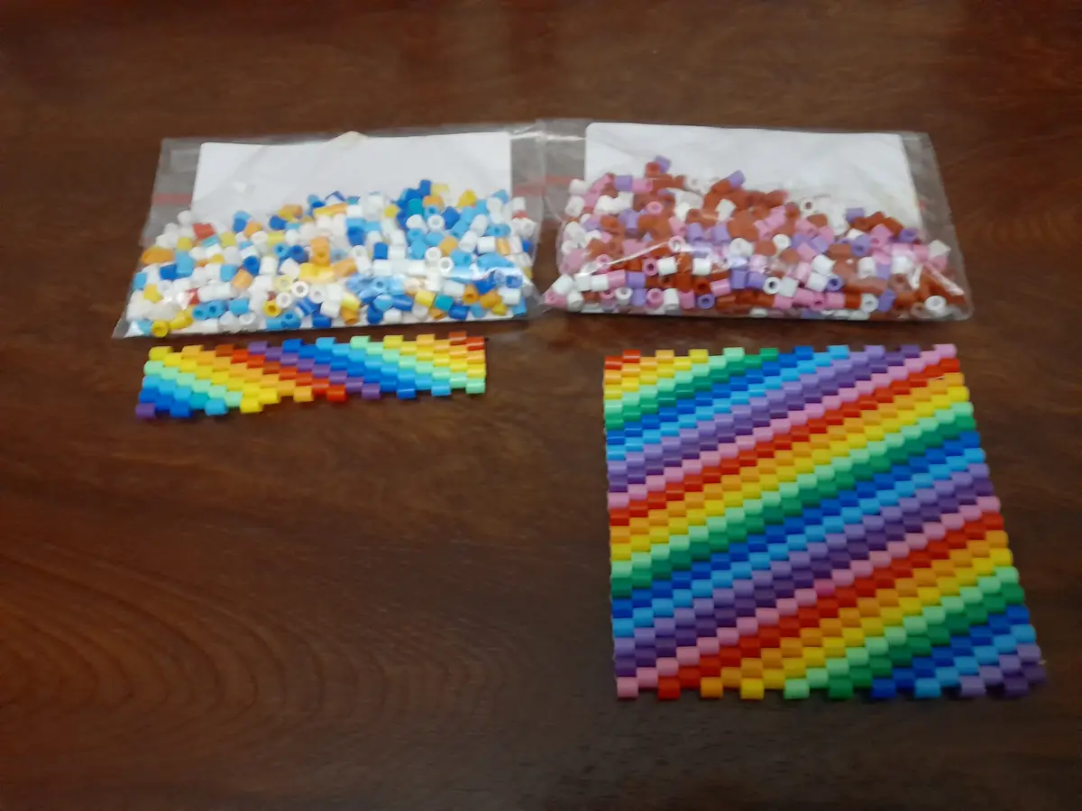 Two clear plastic bags filled with assorted colorful perler beads sit on a wooden table, alongside two completed perler bead…