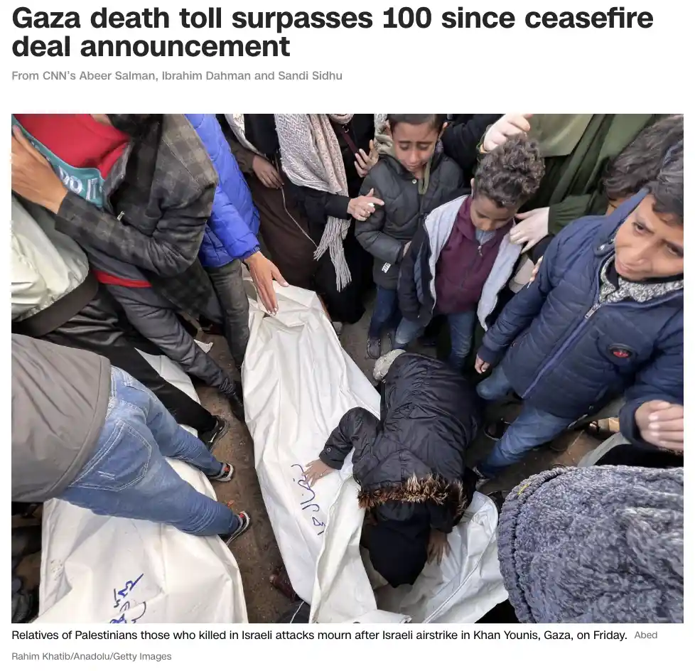 A somber image shows a group of people, including children, gathered around a white body bag with Arabic script, mourning…