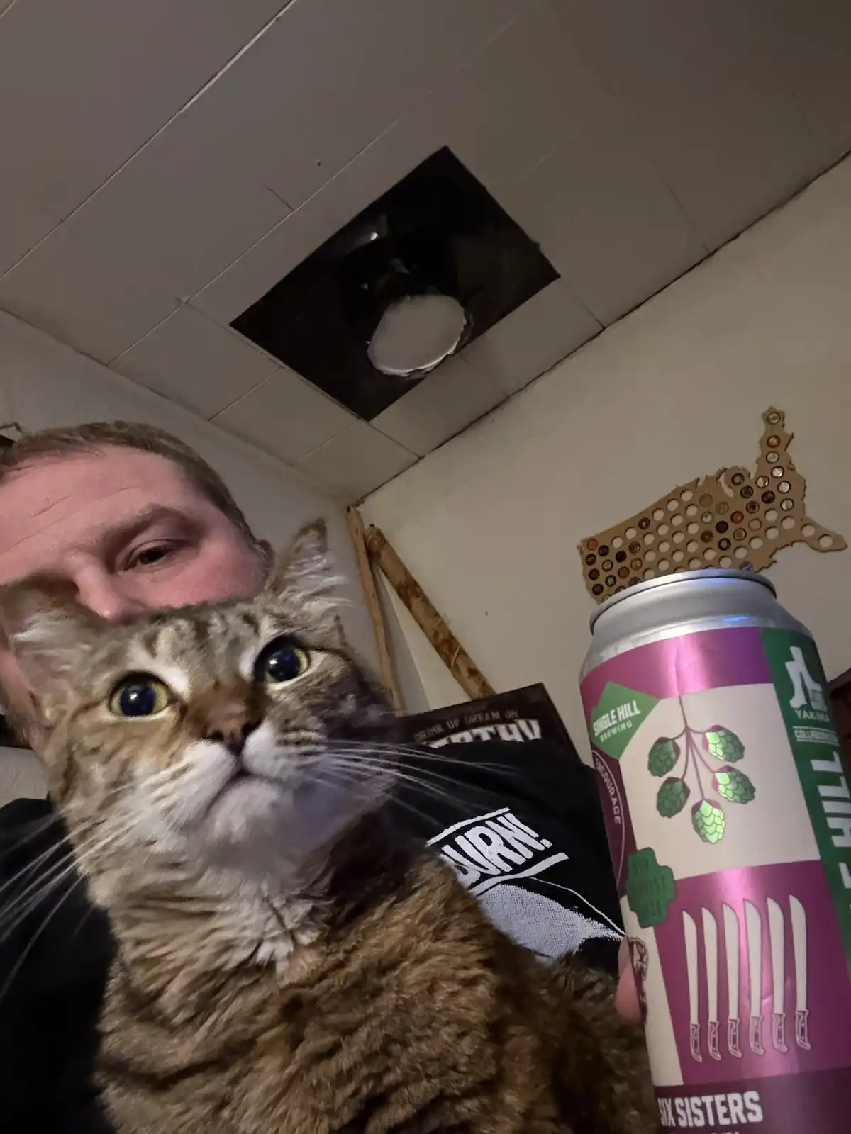 A person holds a tabby cat in front of their face while holding a beer can, with the cat looking directly at the camera with…