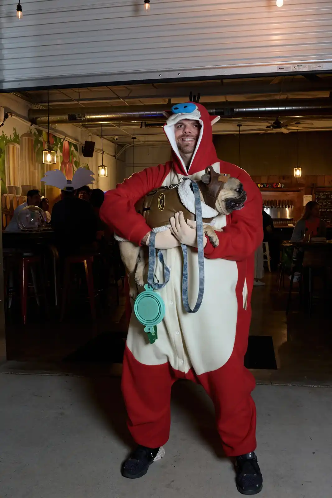 A man in a red and white full-body costume resembling a cartoon character with horns and a blue face holds a pug wearing a…
