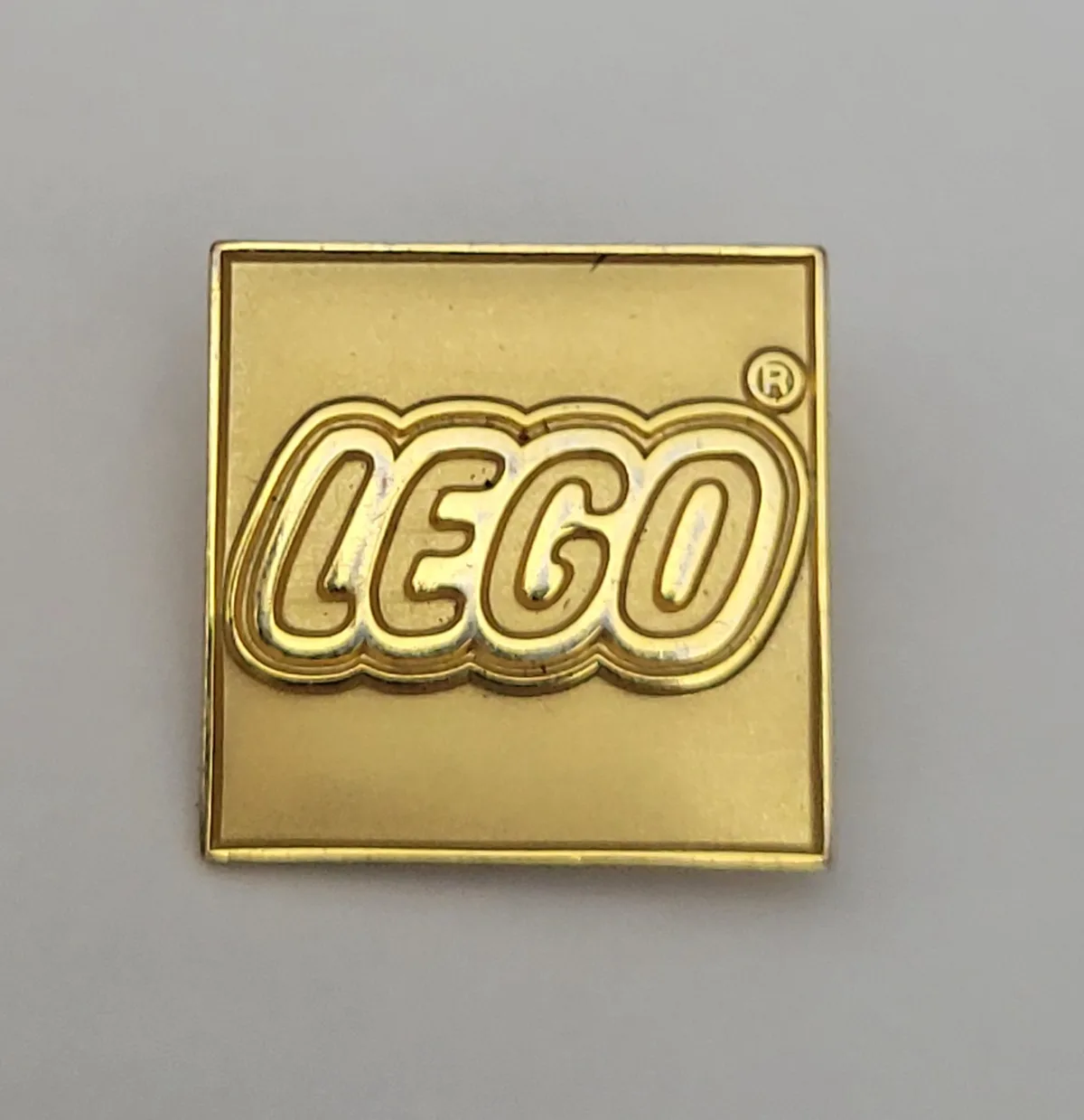 A square, gold-colored pin features the iconic LEGO logo in raised, bubbly lettering with a registered trademark symbol. The…