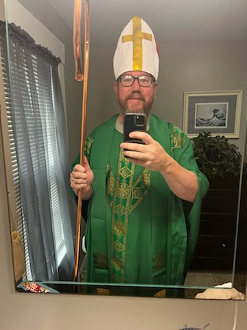 A man in a green and gold clerical robe and a white mitre with a gold cross takes a mirror selfie while holding a staff. He…