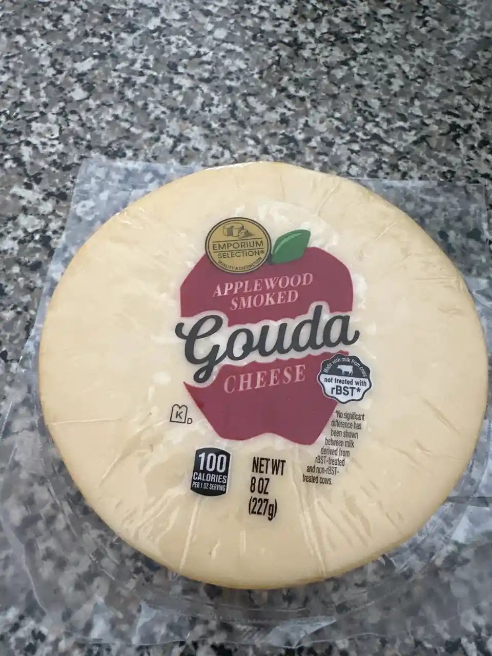 A round wheel of Emporium Selection Applewood Smoked Gouda cheese in clear plastic wrap sits on a speckled gray countertop…