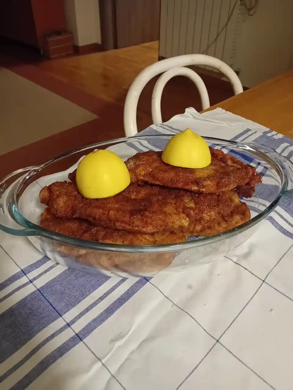 A glass dish holds several golden-brown fried schnitzels stacked on a blue-and-white checkered tablecloth, with two bright…